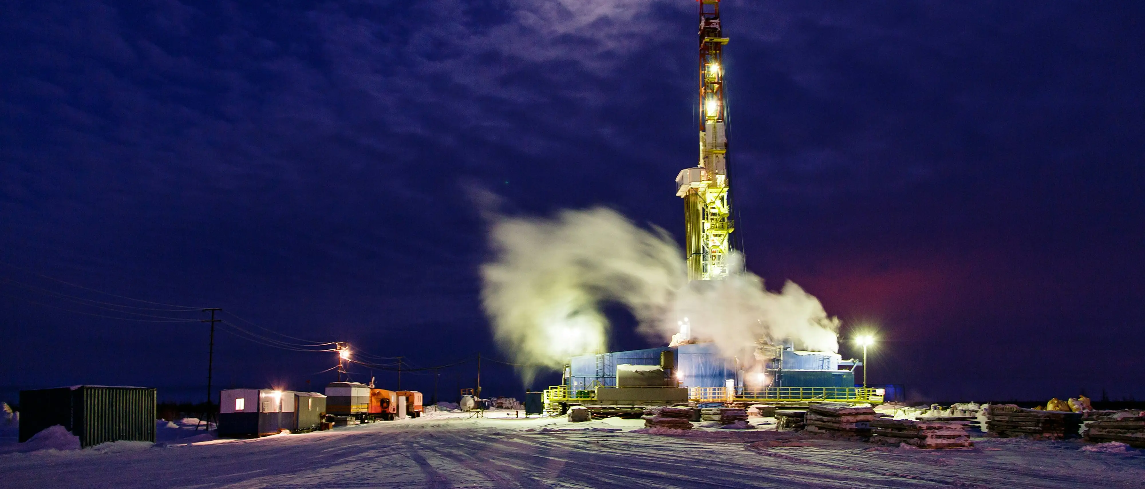 A drilling rig lit up at night on a snow-covered well site under a moonlit winter sky.
