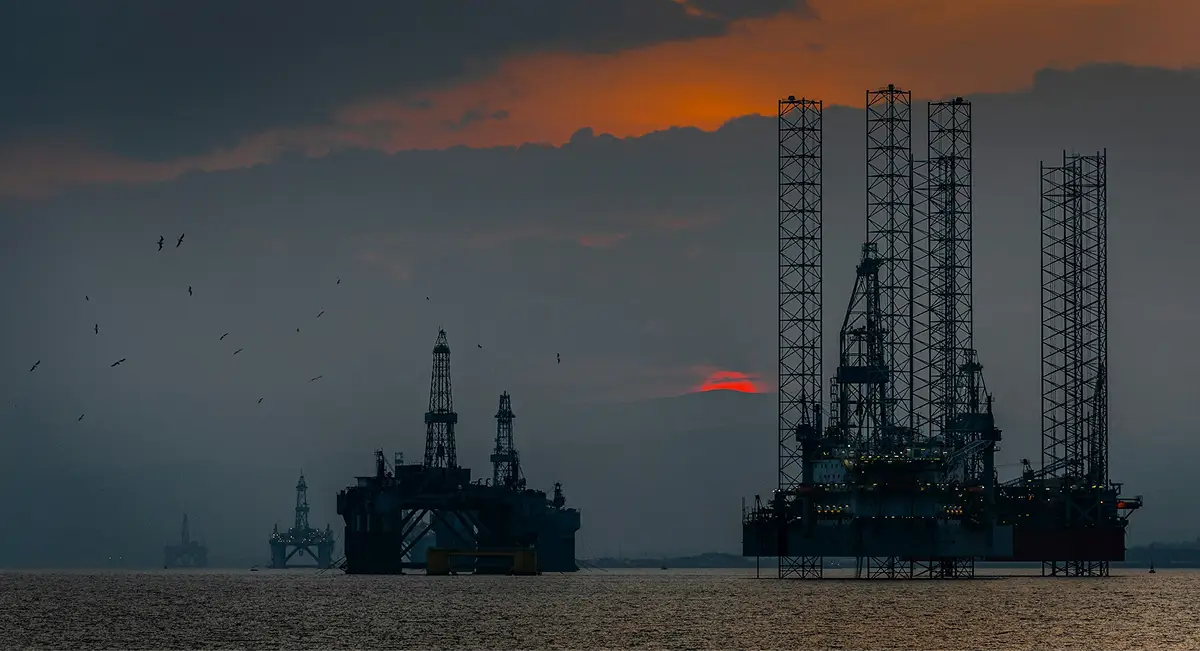 Offshore drilling and production platforms at sea at dusk, with more rigs on the horizon.